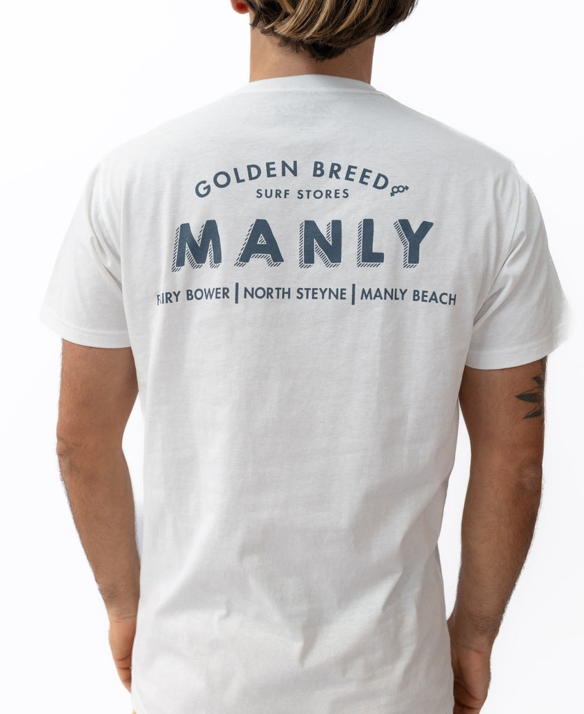 A person wearing the Manly Old School Tee | Off White by Golden Breed—a 100% cotton t-shirt designed in Australia, featuring MANLY Fairy Bower, North Steyne, and Manly Beach printed on the back.