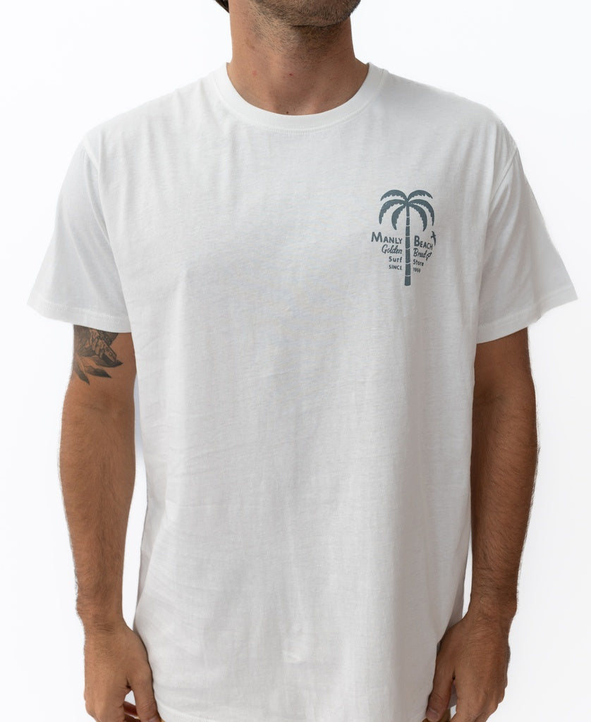 A person wears the Golden Breed Manly Palm Loco Tee in off white, a 100% cotton shirt featuring a small palm tree and Manly Beach Gallery Surf Lounge Bar text on the left chest. Designed in Australia.