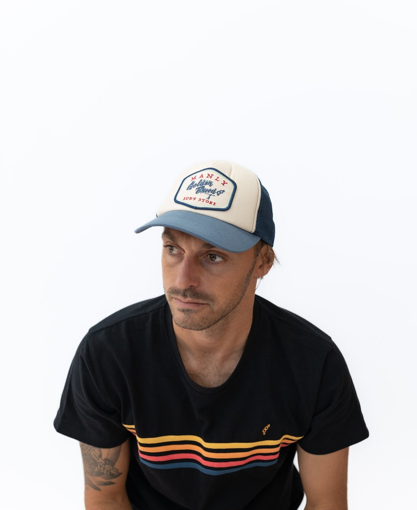 A man wears the Golden Breed Manly Trucker Cap in Dark Denim, featuring an adjustable strap, and a black T-shirt with colored stripes, looking slightly left against a white background.