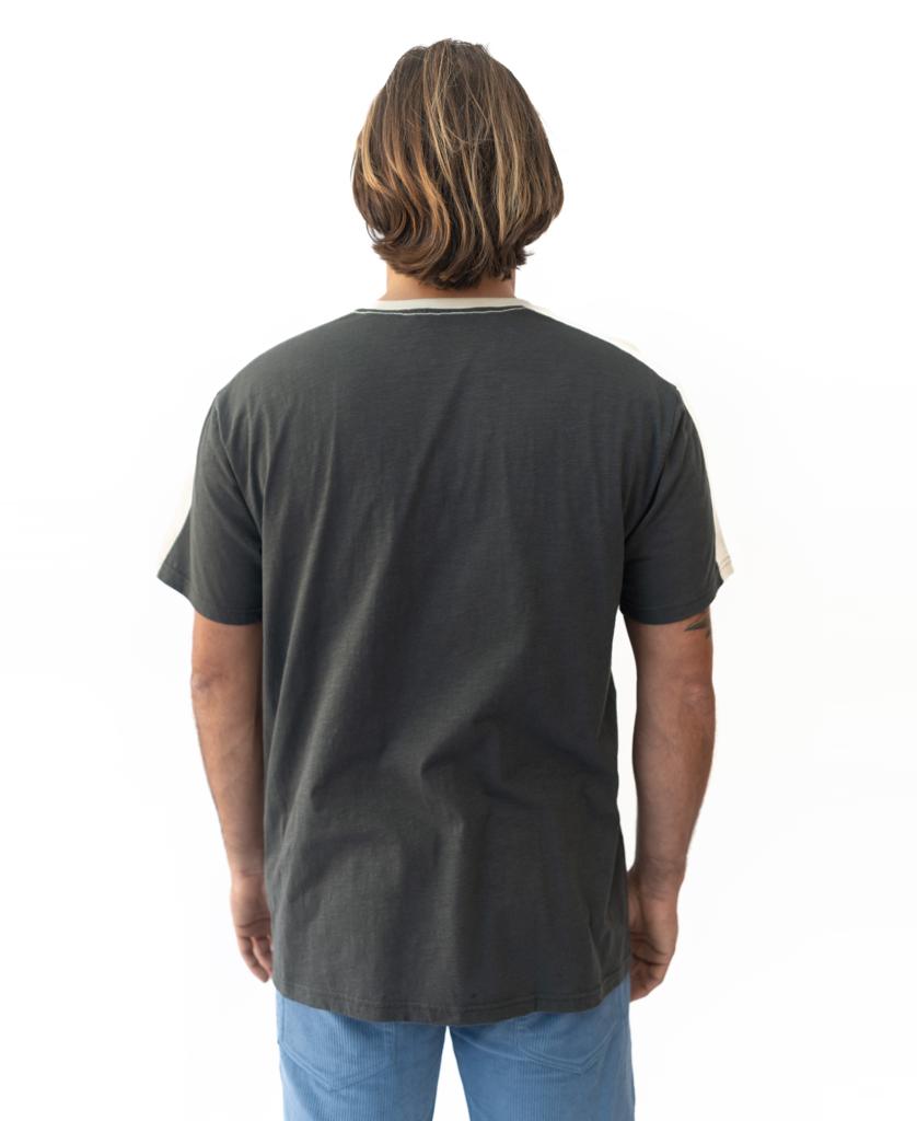 A person with shoulder-length brown hair, wearing a Golden Breed Mitch Tee in Wash Black and light blue pants, stands facing away from the camera against a white background.