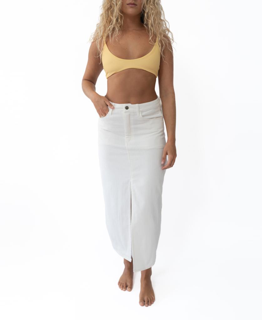 A person with long curly hair wears a yellow bikini top and the Nellie Maxi | Off White skirt by Golden Breed, handmade in Indonesia from soft cord fabric, standing barefoot against a white background.