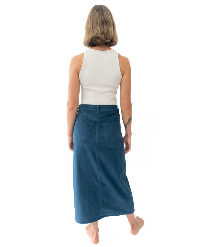A woman stands barefoot, facing away, wearing a white sleeveless top and the Golden Breed Nellie Maxi Skirt in Petrol with back pockets.
