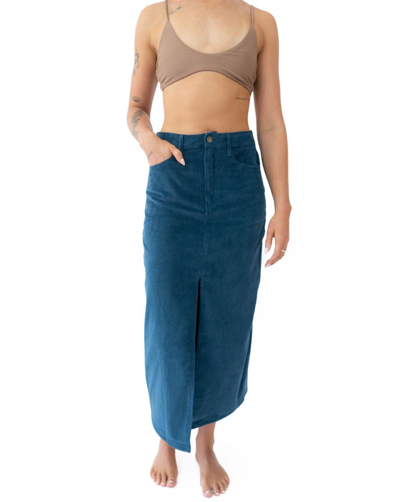 A person stands barefoot against a white background, wearing a tan bralette and the Golden Breed Nellie Maxi Skirt in Petrol—a high-waisted blue maxi skirt with a front slit.