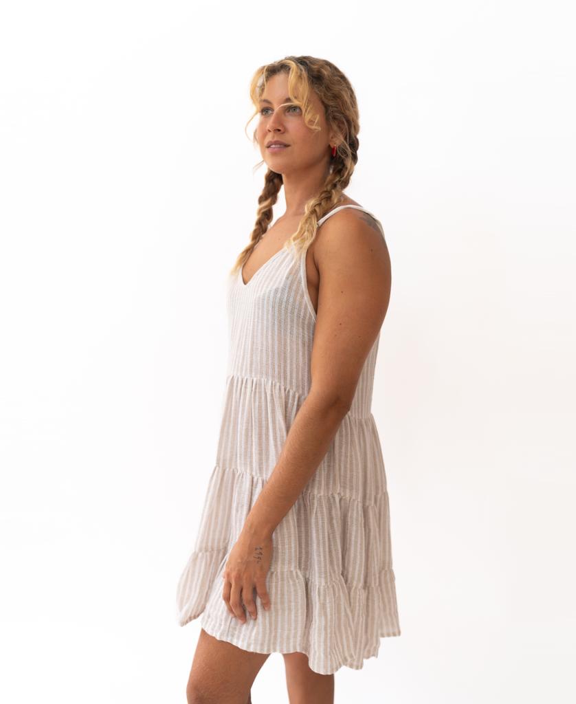 A person with braided hair models the Golden Breed Nina Baby-doll Dress in Off White Stripe, featuring adjustable straps and a sleeveless striped cotton design, while standing against a plain white background facing slightly sideways.