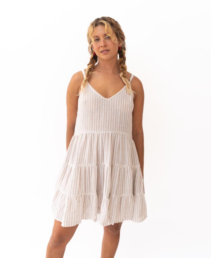 A blonde-haired woman in braids wears the Golden Breed Nina Baby-doll Dress | Off White Stripe, a sleeveless, tiered cotton dress with white and beige stripes, standing against a plain white background.
