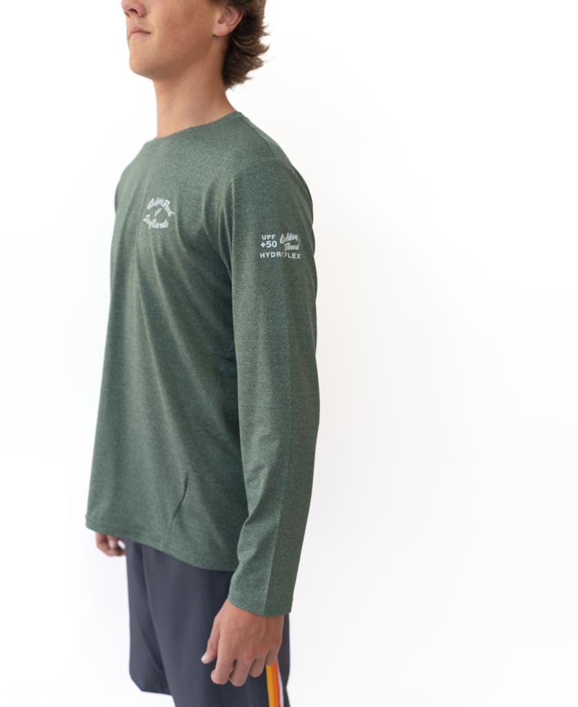 A person wears the Golden Breed Pebbles L/S Rash Shirt in Green Marle, featuring 50+ UPF sun protection and white branding on the chest and sleeve, paired with dark shorts with a colored stripe, standing against a plain white background.