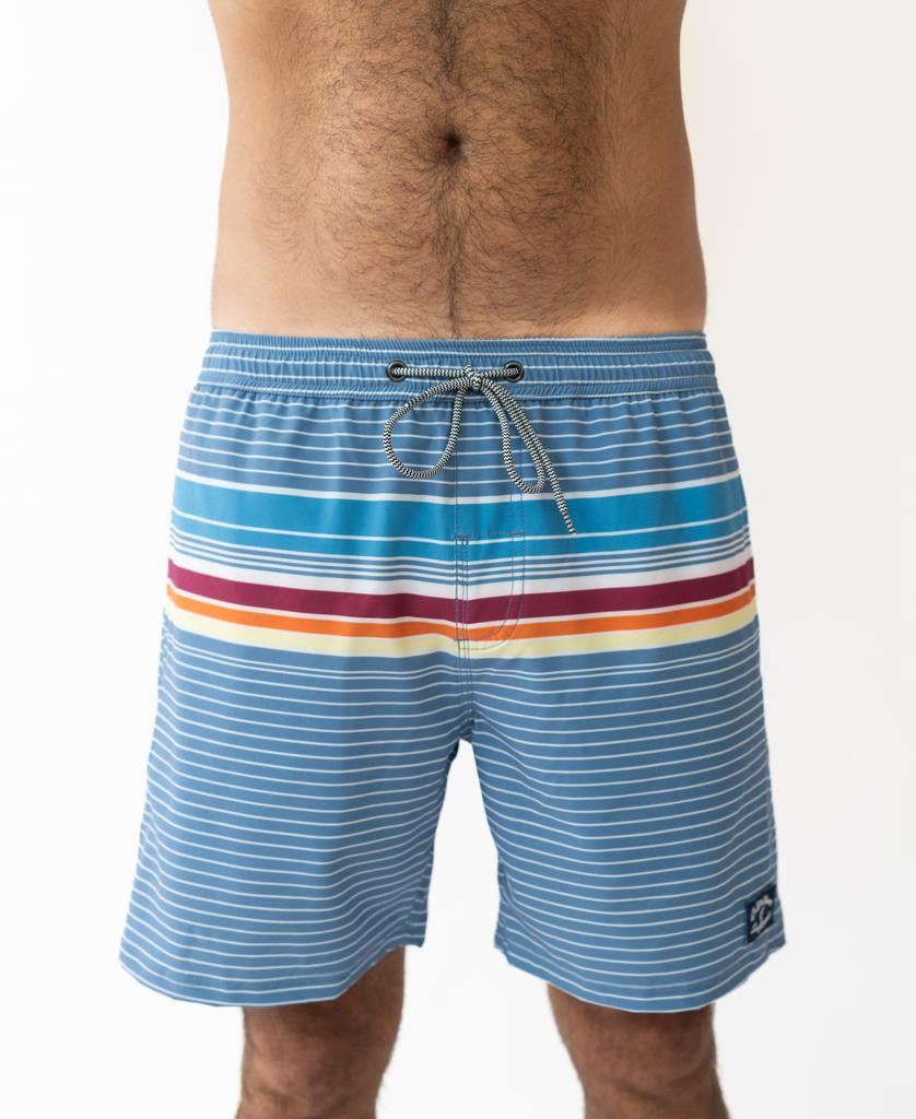A person wearing Golden Breeds Ralph Elastic Waist Boardie in Dusty Blue, featuring blue and white stripes with red and yellow lines, shown waist to mid-thigh. Designed with 4 Way Stretch for comfort.