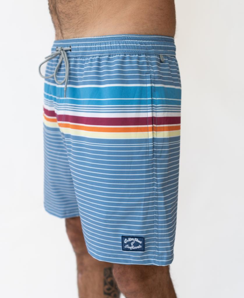 A person is shown from the waist to above the knee wearing the Golden Breed Ralph Elastic Waist Boardie in Dusty Blue, featuring blue stripes, a red and yellow horizontal stripe, an elastic waist, and a drawstring waistband.
