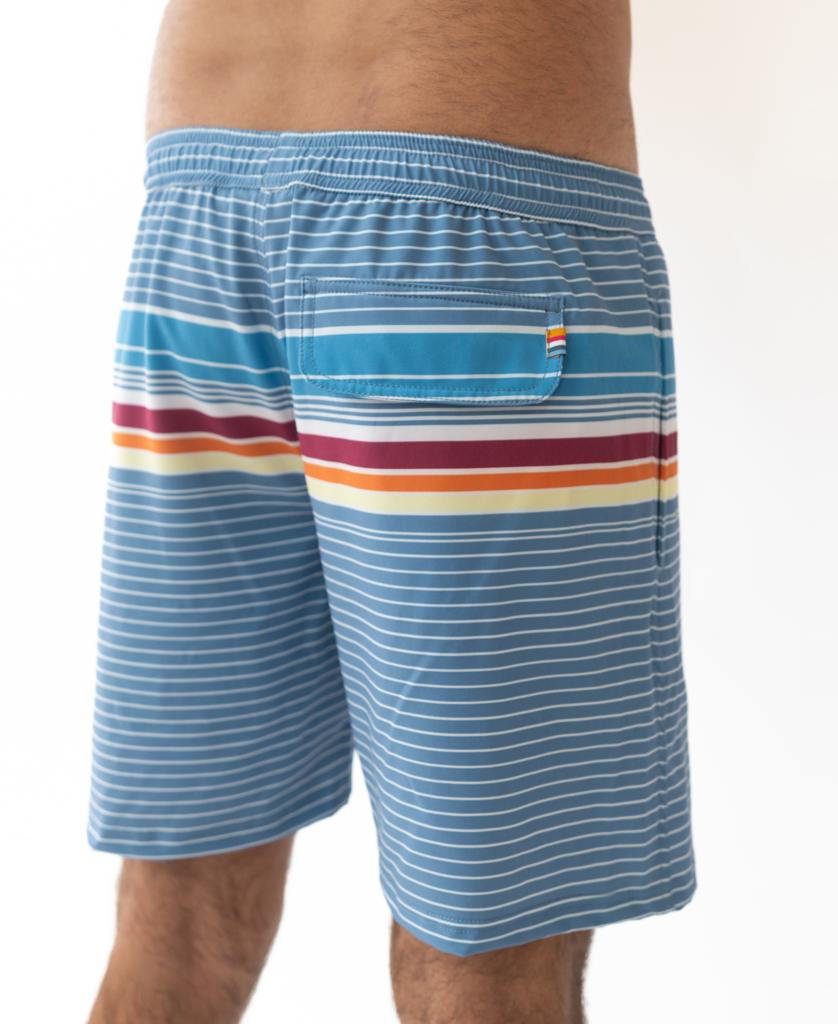 A person wearing Golden Breed’s Ralph Elastic Waist Boardie in Dusty Blue, featuring blue with horizontal red, orange, and yellow stripes and a back pocket, shown from the waist down.