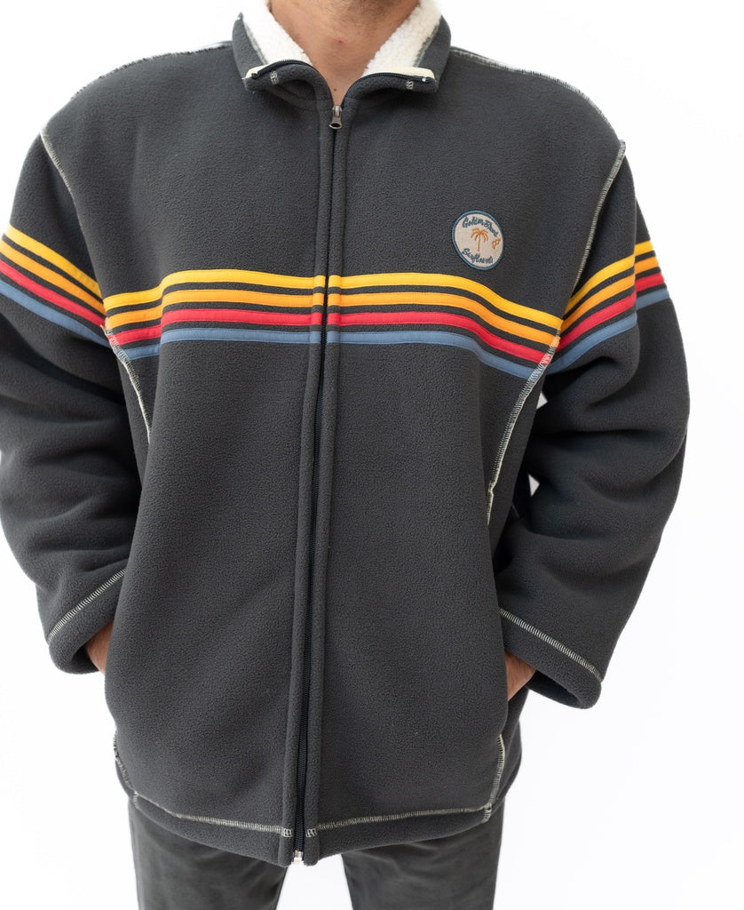 A person wears the Golden Breed Reef Bonded Sherpa Jacket in Wash Black, a dark gray polyester sherpa fleece with red, yellow, and orange horizontal stripes and a small circular chest patch. Their hands are tucked in the jacket pockets.