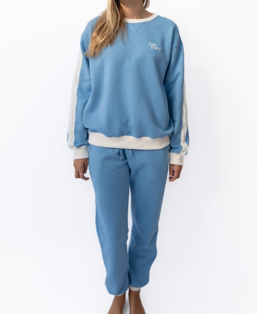 A woman stands against a white background wearing Golden Breed’s Retro Crunchie T/P in Azure/Off White, paired with matching trackpants featuring white sleeve stripes.