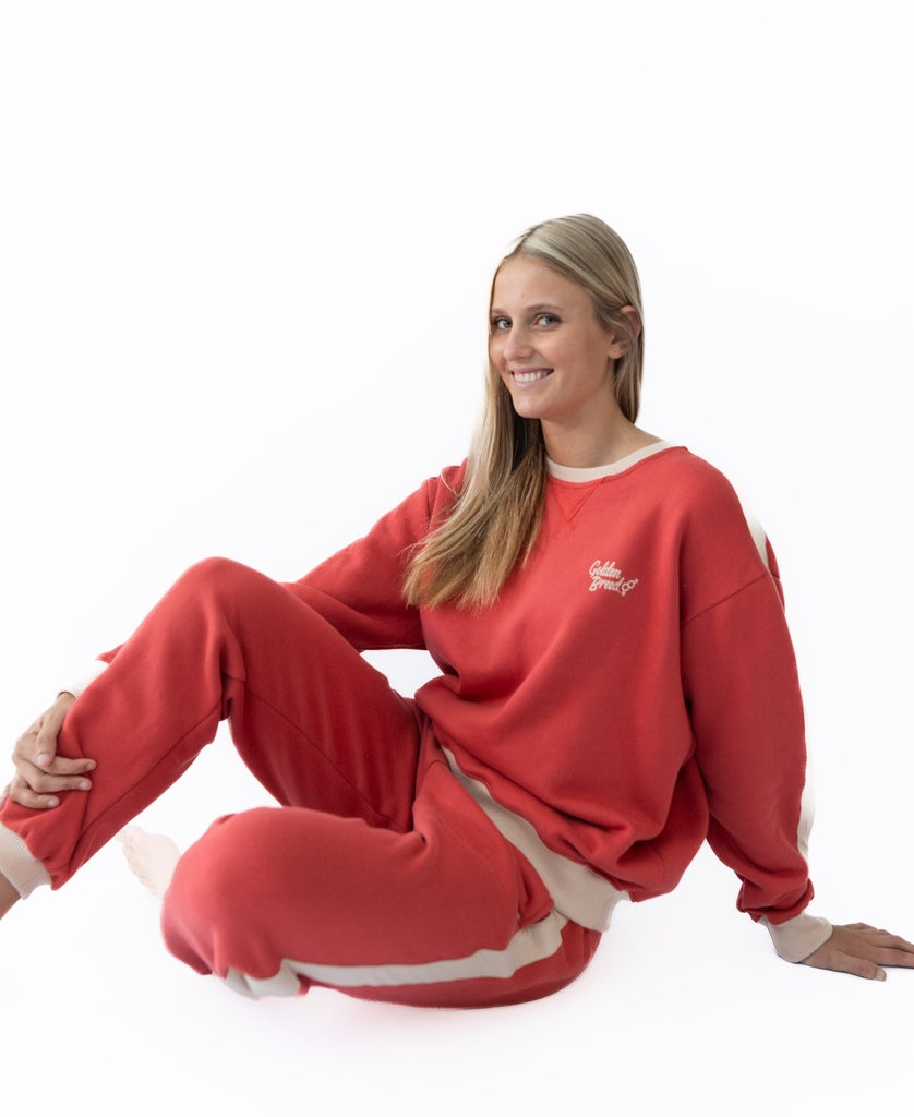 A woman with long blonde hair sits on the floor, smiling at the camera in a Golden Breed Retro Crunchie T/P sweatsuit in wash red/beige, against a plain white background.