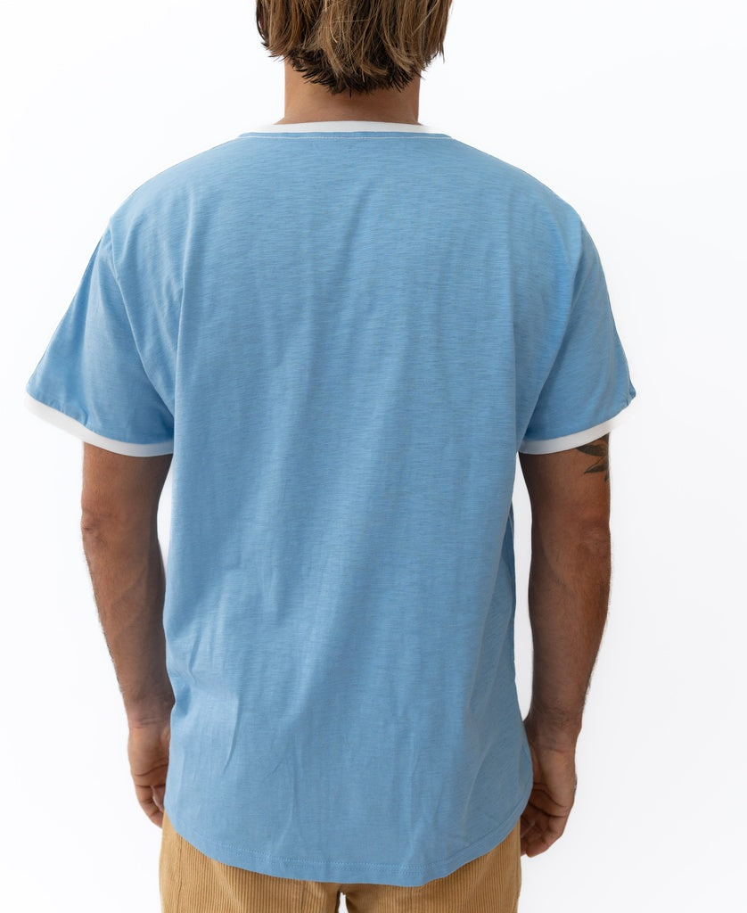 A person stands with their back to the camera, wearing Golden Breed’s Retro Ringer Tee in Azure Blue with white trim and light brown pants, channeling effortless 70s vibes.