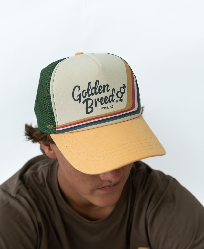 Individual wearing a wash orange Golden Breed Retro Trucker Cap featuring the Since 69 logo and adjustable strap, looking down, paired with a brown t-shirt.