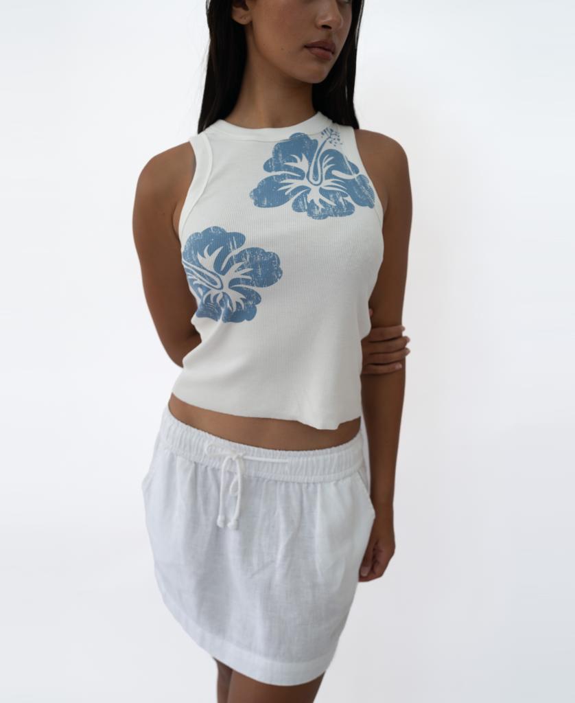 A woman wears the Golden Breed Rosa Hibiscus Tank in Azure—a high neck, white ribbed tank with blue floral prints—paired with a white drawstring skirt, standing against a plain white background.