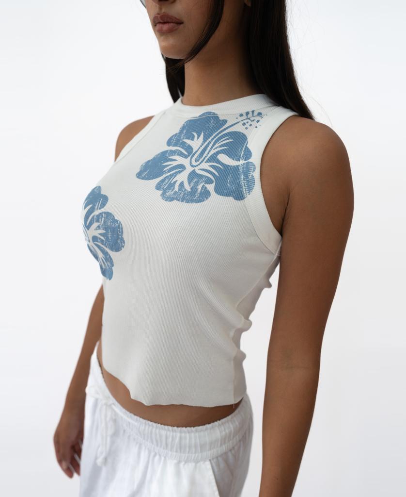 A woman wearing the Golden Breed Rosa Hibiscus Tank in Azure, featuring blue hibiscus prints, and white pants stands against a plain white background.