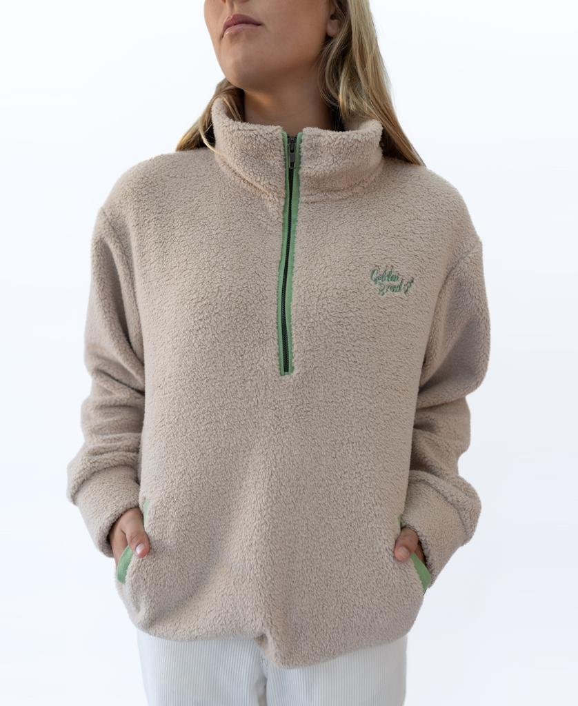 A woman wears the Sammie 1/2 Zip in beige by Golden Breed, featuring a green zipper and Retro Golden Breed logo, hands in pockets, standing against a plain white background.