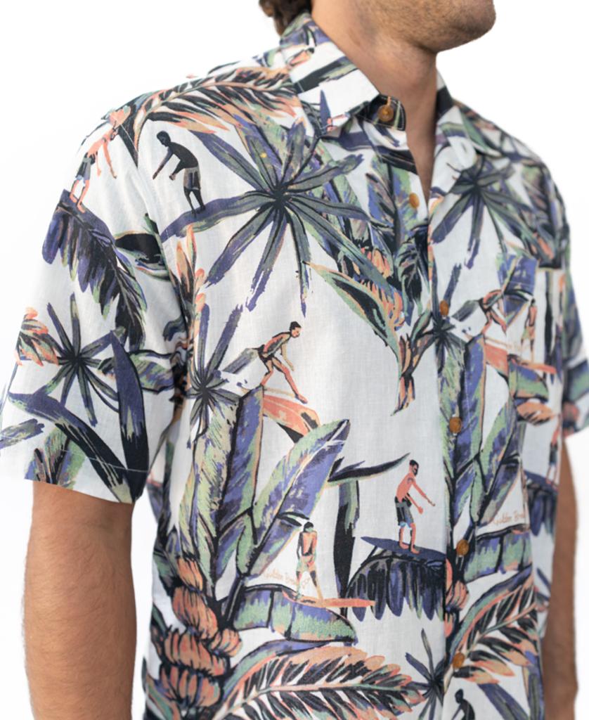 A man wears the Golden Breed Surfer Palm Shirt in Off White, featuring a tropical print with large leaves and surfers, crafted from a comfy cotton/linen blend for laid-back island style.