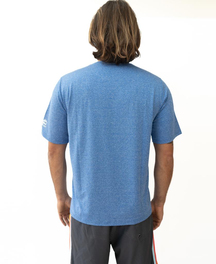 A person with shoulder-length brown hair, wearing a Golden Breed Nemo S/S Rash Shirt in Blue Marle and dark shorts, stands with their back to the camera.