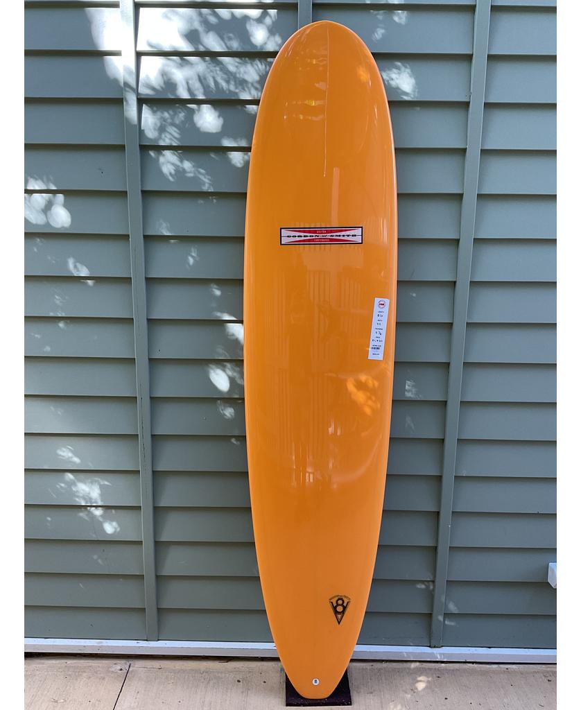 The Gordon & Smith G&S 80 V8 Box & Sides in orange stands upright with its fin against a light green paneled wall on concrete, ideal for small to mid-size waves.
