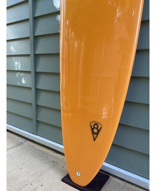 The Gordon & Smith G&S 80 V8 Box & Sides in orange, featuring a black and white logo, stands upright against a light blue paneled wall on concrete—perfect for small to mid-size waves.