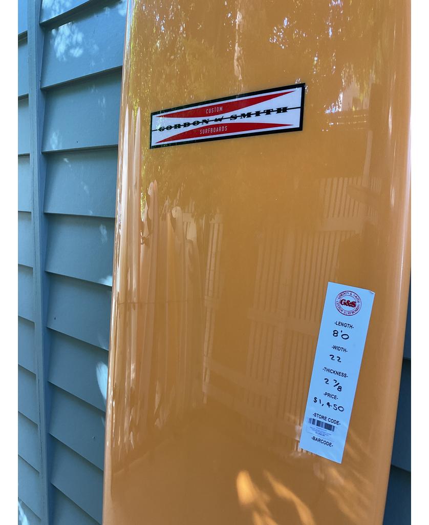 An orange Gordon & Smith G&S 80 V8 Box & Sides mid length board with medium rocker leans against a light blue wall, displaying the logo and details—ideal for riding small to mid size waves.