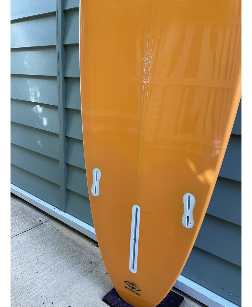 The Gordon & Smith G&S 80 V8 Box & Sides in orange, a mid-length performance board with medium rocker and three fin boxes, stands vertically against a light blue paneled wall on concrete—ideal for small to mid-size waves.
