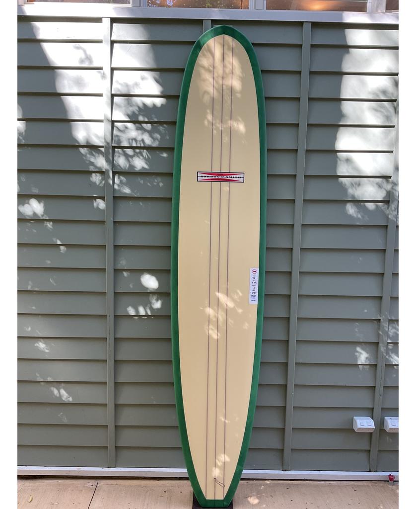 The Gordon & Smith G&S 96 Easy Rider Triple Stringer in Green/Cream, a user-friendly longboard surfboard, stands upright outdoors against a gray-green paneled wall.