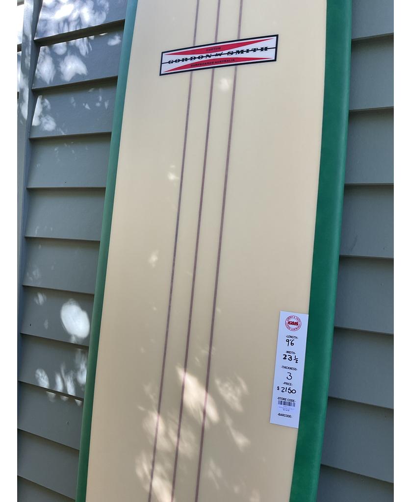 The Gordon & Smith G&S 96 Easy Rider Triple Stringer in green and cream, featuring a medium rocker design, is propped against a gray paneled wall with its specs label attached near the bottom.