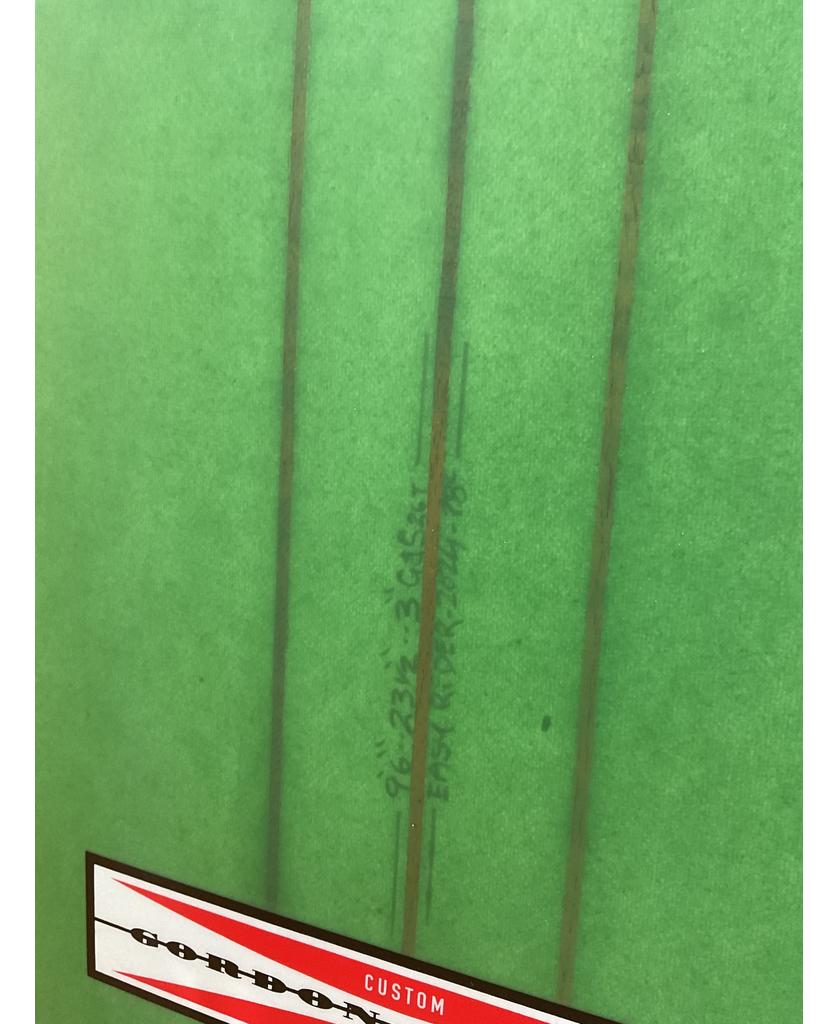 A close-up of the Gordon & Smith G&S 96 Easy Rider Triple Stringer longboard in green/cream, featuring three thin brown stringers, handwritten numbers, and a partial red-and-white striped custom label in the bottom left corner.