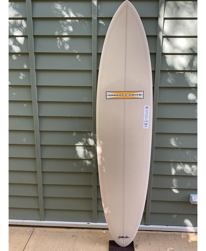 A G&S 78 Cool Tool Mid Twin surfboard in cream by Gordon & Smith stands upright against a green paneled wall, sunlight highlighting its design and the label on the upper half.