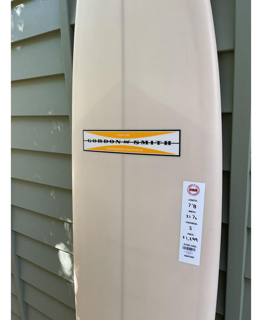 A cream Gordon & Smith G&S 78 Cool Tool Mid Twin surfboard stands upright against a gray wall, displaying its brand label and sales tag with dimensions and price, ready to elevate your surfing experience.