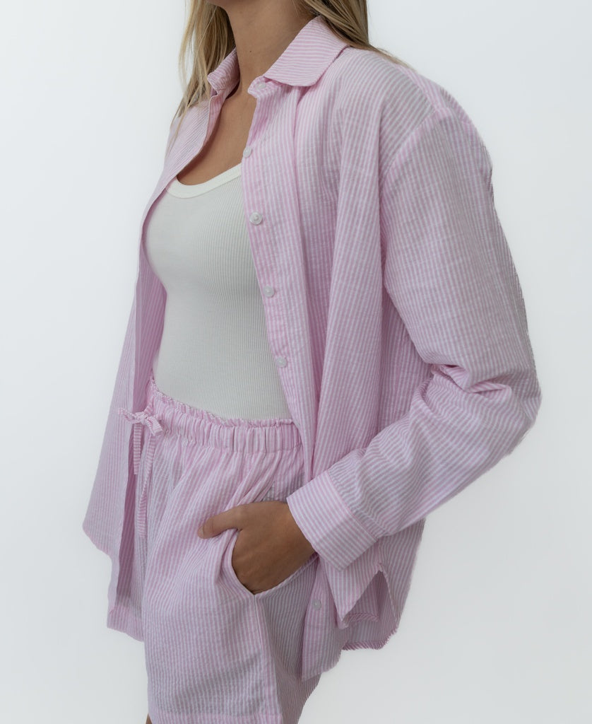 Wearing the Lazy Beach Maeve L/S Shirt in Baby Pink Stripe over a white tank and matching shorts, the person stands with one hand in their pocket against a plain background.