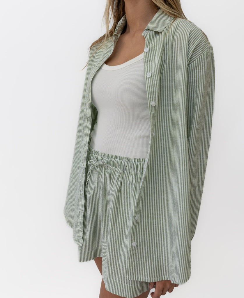 A person wears a white tank top styled with the Lazy Beach Maeve L/S Shirt in Olive Stripe and matching high-waisted shorts.