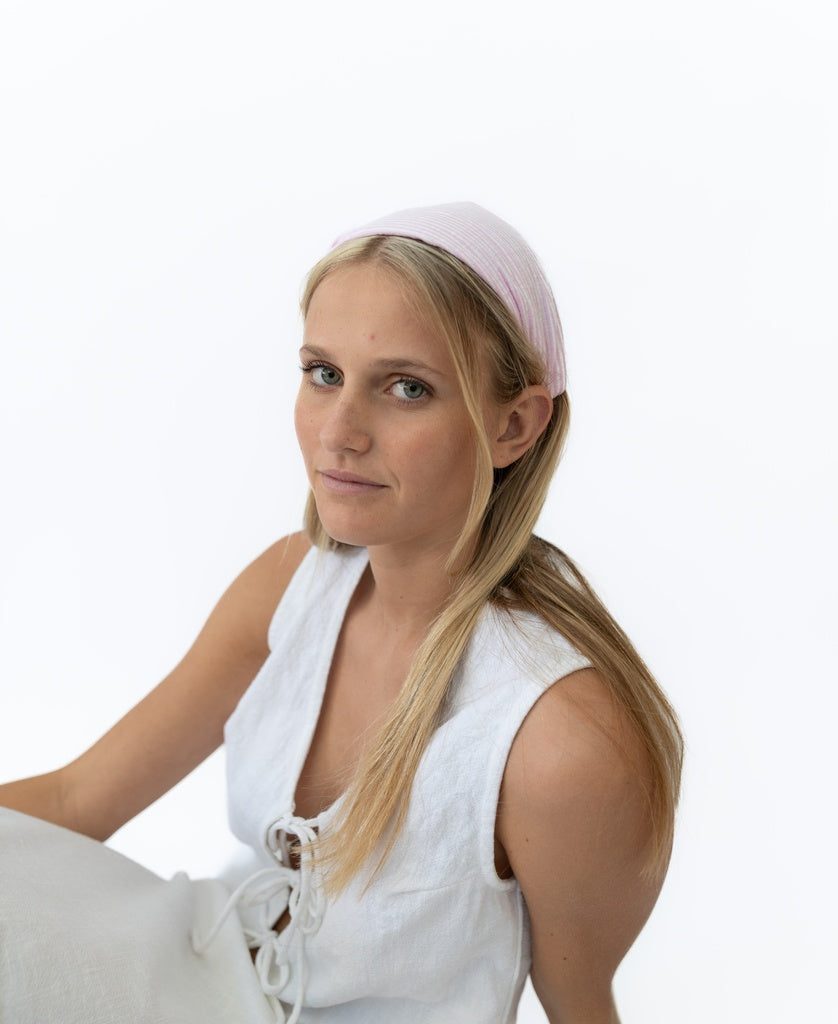 A woman with long blonde hair wears the Lazy Beach Head Scarf in Pink Stripe, handmade in Bali, paired with a white sleeveless top against a plain white background.