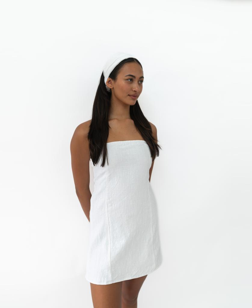 A woman with long dark hair wears the Lazy Beach Head Scarf in White and a strapless white dress, standing against a plain white background with her hands behind her back.
