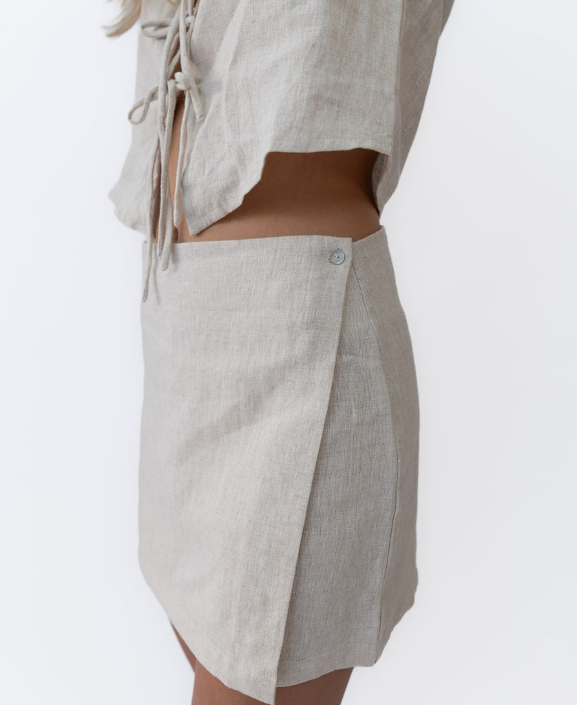 Someone wearing Lazy Beach’s Indiana Skort Linen in Natural—a light beige, 100% linen high-waisted skort with side buttons—paired with a matching short-sleeve tie-front top.