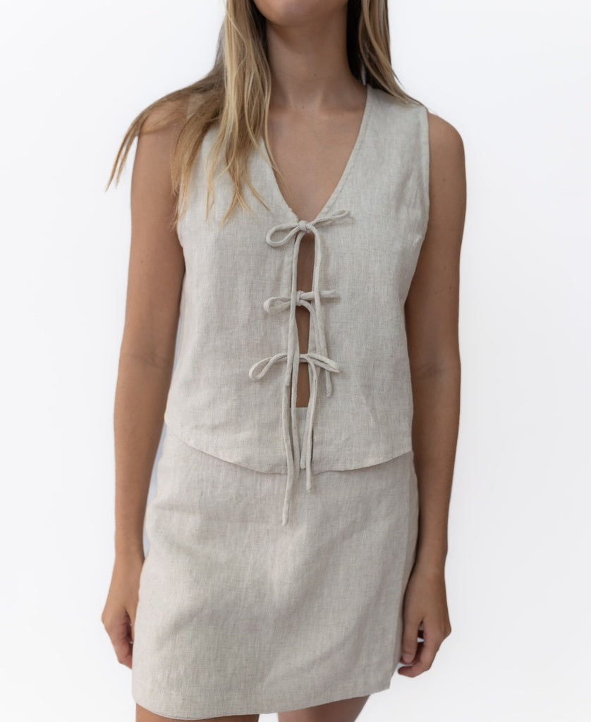 A woman wears Lazy Beach’s Indiana Skort Linen in Natural with a matching sleeveless, light beige 100% linen top featuring three front tie closures, standing against a plain white background.