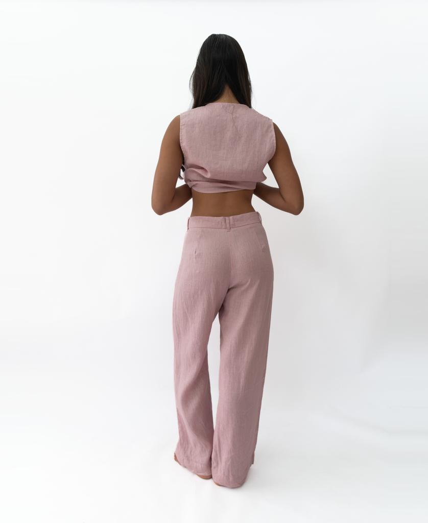 A person stands facing away from the camera, wearing a sleeveless mauve crop top and Lazy Beach’s Janine Pant in Linen Blush, set against a plain white background.