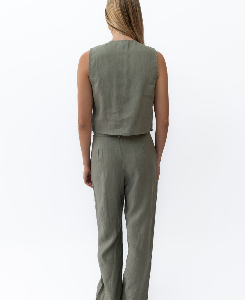 A person faces away, wearing the Lazy Beach Janine Pant Linen in dark khaki with a sleeveless, light olive green top against a plain white background.