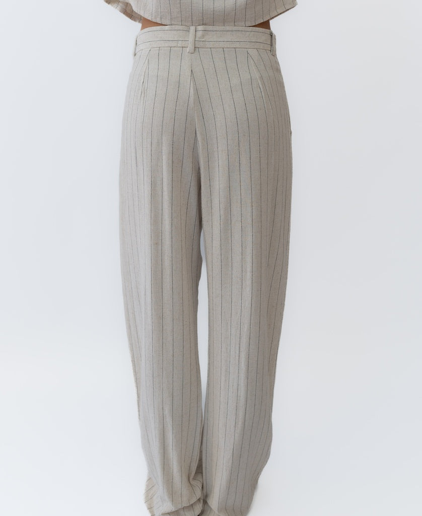 A person wearing Lazy Beachs Janine Pant | Fine Stripe—a high-waisted, wide-leg beige linen rayon blend with front pleats—stands against a plain white background.