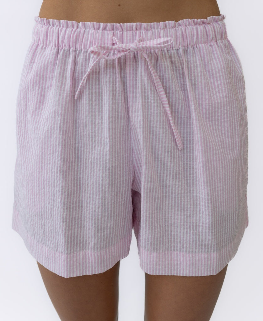 A person models the Judith Short | Baby Pink Stripe by Lazy Beach, handmade cotton seersucker pajama shorts with light pink and white stripes and a drawstring waist, shown from waist to mid-thigh.