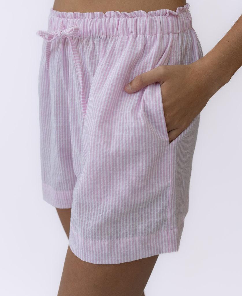 A person wears Lazy Beachs Judith Short in baby pink stripe, handmade from white cotton seersucker with an elastic waistband and drawstring, shown from the waist down with one hand in the pocket.