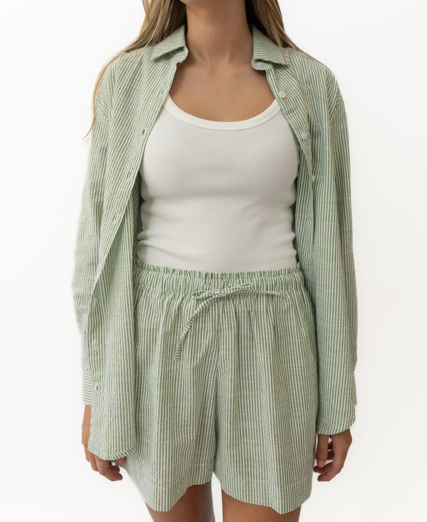 A person wearing Lazy Beach’s Judith Short in Olive Stripe—elastic waist, handmade in Indonesia—with a matching button-up shirt and white ribbed tank top.