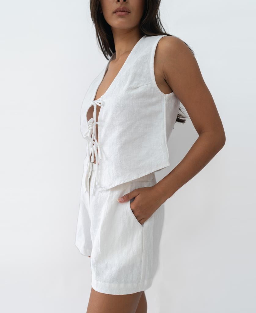 A woman models the Kris Short in White by Lazy Beach—a sleeveless linen vest with front ties and matching mid-length shorts, handmade in Bali—posing against a plain white background for a chic summer vibe.