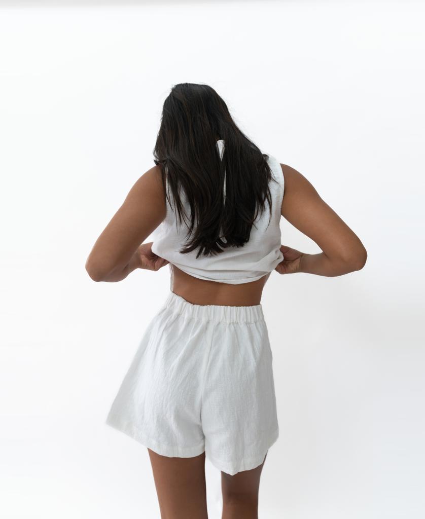 Seen from behind against a plain white background, a person with long dark hair adjusts their Kris Short in White by Lazy Beach, styled with a sleeveless top for a chic summer look.
