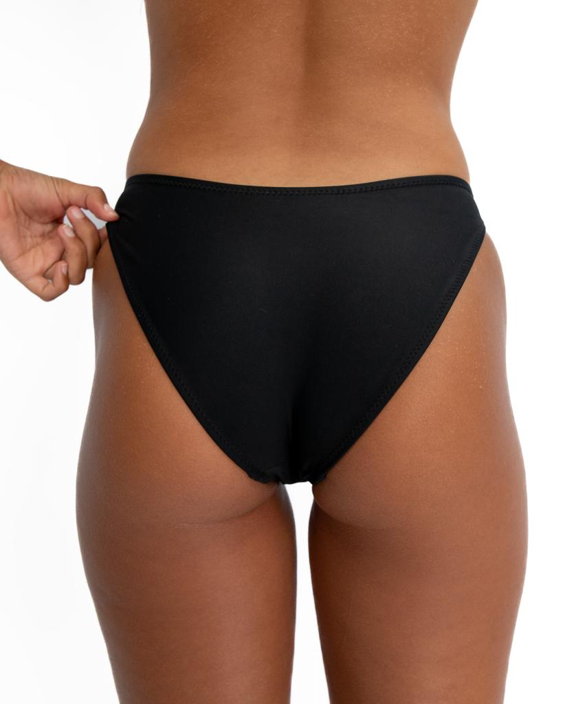 A person wears Lazy Beach Millie Bottom | Black Textured bikini bottoms, shown from the back against a white background, with one hand adjusting the waistband.