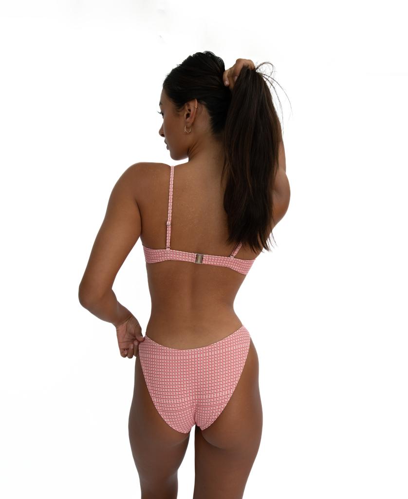 A woman stands against a white background, facing away, wearing Lazy Beach’s eco-friendly Mischa Bikini Top in Pink Houndstooth, made from recycled nylon, and holds her hair up with one hand.