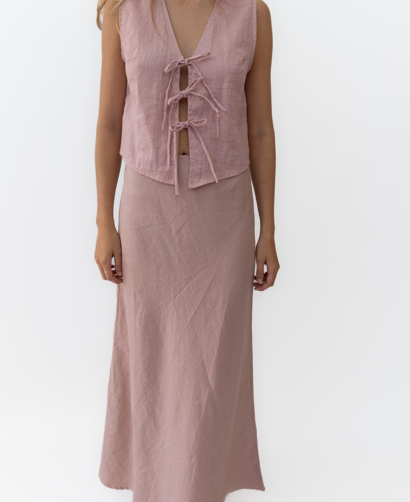 A person wears a sleeveless light pink top with three front ties and a matching Lazy Beach Pearl Maxi Skirt Linen in Blush, standing against a plain white background.