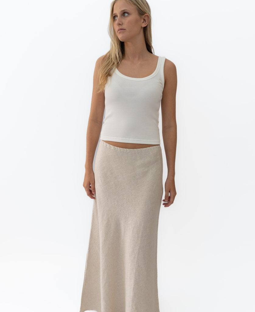 A woman wears Lazy Beachs Pearl Maxi Skirt Linen in natural linen with a sleeveless white tank top, standing against a plain white background.